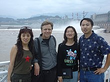 Ted Conover stands with two Chinese women and one Chinese man. Behind them are the Yangtze River, the Three Gorges Dam, several large construction cranes, and misty hills. Buildings dot the area around the base of the hills.