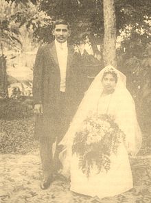 A faded sepia-toned wedding portrait from Sri Lanka; the groom is standing in a long frockcoat; the bride is seated in a white gown and veil, with a large bouquet on her lap.