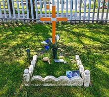 The image shows an orange wooden cross, in front of which is a small 0.5m square grass area bounded by stones, with some flowers and a small photograph of Karl.
