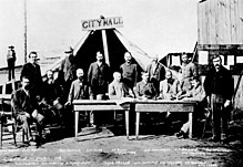 Men standing and sitting around two tables, facing the camera. A large tent behind them has a wooden sign that reads "City Hall"