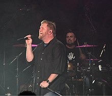 In a dark room, a singer holding a microphone in one hand and stand in the other is standing in front of a drummer