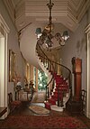 Indoor spiral starcase with red carpeting next to a grandfather clock and a chandelier