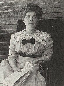 A middle-aged Emma Wolf sitting on a chair, holding a newspaper