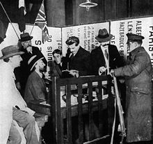 Photo en noir et blanc. Derrière une table-bureau abritée par une barrière en bois se pressent six hommes affairés en complet veston, pour la plupart chapeautés ; devant la barrière un soldat anglais reçoit un ticket de repas (il « est vu acceptant un ticket » dit la légende en anglais. La scène se passe dans un angle réduit formé de panneaux d’avis et donne une impression de confusion.