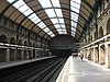 A railway station with side platforms either side of two tracks that disappear into darkness under a painted steel bridge like structure topped with a brick wall, covered by a partially glazed barrel roof.