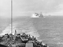 Black and white photo with part of a ship in the foreground and flames and smoke risking from the left of a warship in the background. Several other ships are visible on the horizon at the rear of the photo.