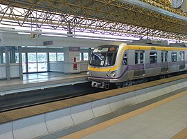 A squarish aerodynamically contoured front of a metro train with purple and yellow stripes running along its sides enters from the right into an empty station that is lit up in part by sunlight streaming in from glass doors and windows.