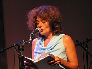 Mishori, reading her poetry. She is standing before a mic, holding a book in both hands.