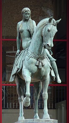 The statue from the front. It is made of bronze, now green, and depicts Lady Godiva sat sidesaddle on a horse. She is naked, with her long hair falling over her left breast. The horse (from its perspective) has its front right hoof raised and its head points left. The building behind has a facade of black glass in red frames.