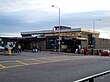 A building with a blue sign reading "BLACKHORSE ROAD STATION" in white letters and several people and cars in the foreground all under a blue sky