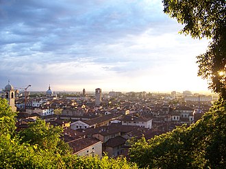 The sun, low on the horizon, bathes a small city in light; the light casts deep shadows on tiled roofs, church towers, gardens, and walls.