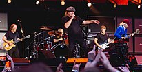 Five men perform on stage: The rhythm guitarist looking at his guitar, drummer behind his kit, lead singer in the middle, bass guitarist turned to his right and lead guitarist in school boy costume.