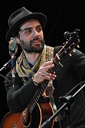 Oscar Isaac is holding a guitar while looking to his right.