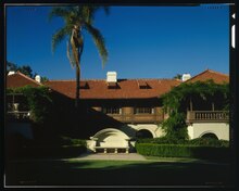 Building with terra cotta roof, a palm tree to the left and well trimmed hedges on the right with a cloudless sky