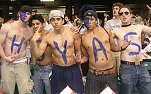 Five young shirtless men pose defiantly in a crowd. Each has a letter in blue on their chests to spell HOYAS.