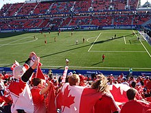 Photo d'un terrain pendant un match prise d'une tribune latérale. Au centre un fan porte le drapeau du Canada sur son dos.