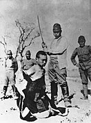 Photographie en noir et blanc d'un homme agenouillé au premier plan, le cou et les épaules dénudés. En arrière-plan un homme portant un uniforme s'apprête à porter un coup de sabre sous le regard de trois autres personnes.