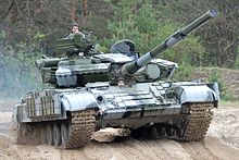 Vor einem Wald steht ein Panzer mit zwei Soldaten, die aus ihren Ausstiegsöffnungen blicken.