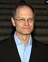 A man in his early fifties looking to the left of the camera. He is wearing thin rectangular glasses and a black blazer with a light-colored shirt.