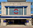 A grey building with a rectangular, dark blue sign reading "MORDEN STATION" in white letters, two red buses in front, and a fence in the foreground