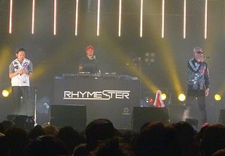 Three Japanese hip hop artists on a stage, one holding a mic up to his face.