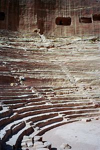 Vue en perspective des gradins d'un théâtre romain avec un âne sur l'un d'eux. En arrière plan des ouvertures approximativement rectangulaires sont pratiquées dans la roche.