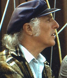 blurry close-up image of Gilles Vigneault from profile, wearing a white shirt, brown plaid jacket, and blue brimmed hat, squinting with his mouth partly open