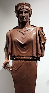 Red stone statue of a woman, looking straight ahead and tugging at her clothing with her right hand.