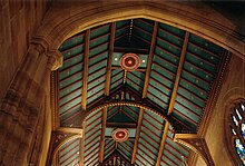 View looking up into the open gabled roof of St Andrews. The beams, rafters and air vents are decorated with bright red rebates and gold leaf details. The ceiling panelling is blue-green dotted with gold stars.