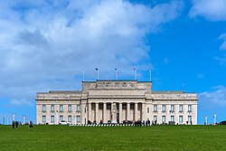 Large neoclassical-style building with a forecourt featuring a Cenotaph on a Court of Honour. Above the front porch of the building is inscribed a funeral oration attributed to the Greek General Pericles, which reads "MCMXIV – MCMXVIII / The whole earth is the sepulchre of famous men / They are commemorated not only by columns and inscriptions in their own country / but in foreign lands also; by memorials graven not on stone / but on the hearts of men." A New Zealand flag atop the building is flown at half-mast. Banners hanging between the columns advertise exhibitions about volcanoes, and Charles Darwin.