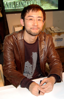 A black-haired man in his forties, wearing a brown leather jacket, sits at a desk at a press event.