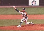 A man in a white baseball uniform with orange and red stripes across the chest throws a baseball with his right hand from a dirt mound on a grass field.