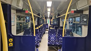 Photo taken inside the train - there are rows of seats on both sides. One side has two seats, and other side has three seats. The seats are blue with multi-coloured spots all over. There are yellow handles for support along the way.