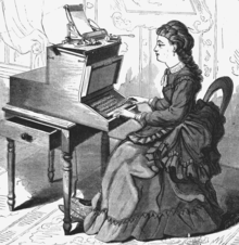 A woman in a long, frilled dress with a pleated train sits at a large, wooden typewriter which is approximately half her size. The typewriter is affixed to a small table with an open drawer.