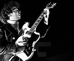 black-and-white image of Yonatan Gat from profile, wearing a turtleneck sweater and dark jacket, playing an electric guitar