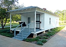 Present-day photograph of a whitewashed house, about 15 feet wide. Four bannistered steps in the foreground lead up to a roofed porch that holds a swing wide enough for two. The front of the house has a door and a single-paned window. The visible side of the house, about 30 feet long, has double-paned windows.