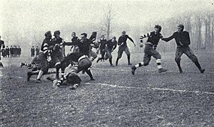 A group of men are playing football. One man is carrying a football, and several others are in his immediate surroundings; six or seven other men are running toward him. A small group of onlookers are watching in the background.