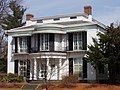 A white two story house with black shutters. Windows on both floors extend from floor to ceiling. The central portion of the house has a protruding rounded porch, supported by white fluted pillars below, and decorated on the second floor with wrought iron railings.