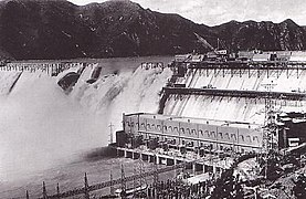 Photographie d'un barrage vu de loin. Des trombes d'eau s'échappent par le sommet du barrage. Au premier plan, un grand bâtiment de plusieurs étages est accolé à l'édifice.