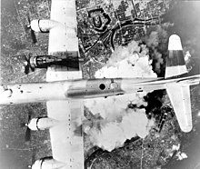 Black and white photo of a four engined World War II-era aircraft being viewed from above while it is flying over a city. A large cloud of smoke is visible immediately below the aircraft.