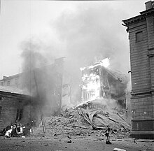 An apartment building is on fire and has partly collapsed in Central Helsinki after Soviet aerial bombing on 30 November 1939. A woman in a coat and a hat is passing by on the right next to the rubble and a car is on fire on the left.