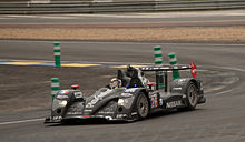 An black open cockpit LMP2 sports prototype car being driven around on a motor racing track