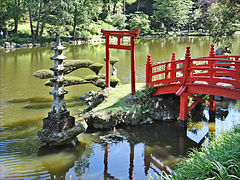 Photo d'un jardin ; un étang central est bordé par une nuée d'arbres ; au premier plan, un pont à la japonaise permet de relier un îlot.