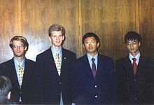 Four boys, in their late teens, wearing shirts, ties and blazers, standing in a line. The two on the left are white, while the two on the right are of East Asian heritage