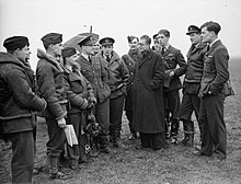 Photographie en noir et blanc représentant un groupe d'hommes se tenant sur un aérodrome. À l'arrière plan, on aperçoit l'aile d'un avion de chasse.