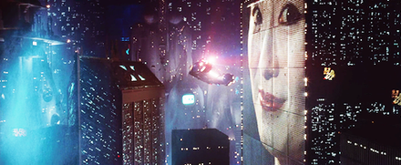 A still frame from the film Blade Runner. A flying car flies between skyscrapers in a futuristic looking city. The buildings are covered with lights and advertising video billboards.