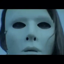 The face of a woman wearing a white mask. Black bars appear at the top and the bottom due to letterboxing.