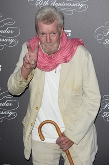 Robert Freeman in front of grey banner holding a cane and giving a peace sign.