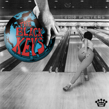 A black and white photograph of a bowling alley, with a hand holding a blue bowling ball with the band's name on it in the top-left.