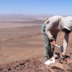 researcher bends over using rock hammer with desert in background